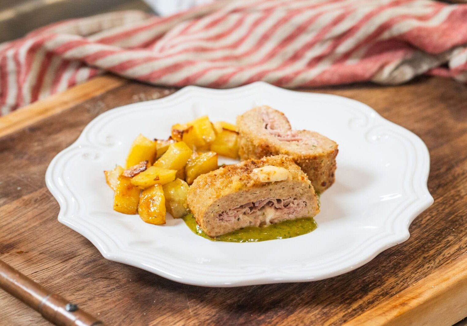 A white plate with breaded ham and cheese rolls sliced in half, served with roasted potatoes and a green sauce on a wooden surface.