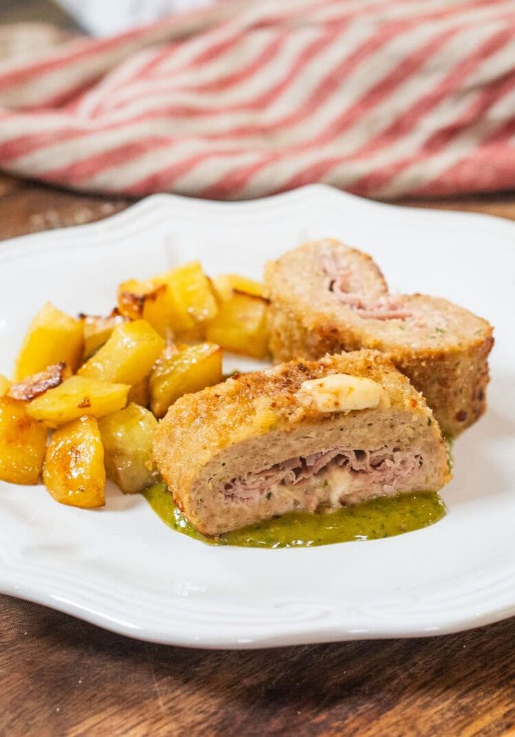 A white plate with breaded ham and cheese rolls sliced in half, served with roasted potatoes and a green sauce on a wooden surface.
