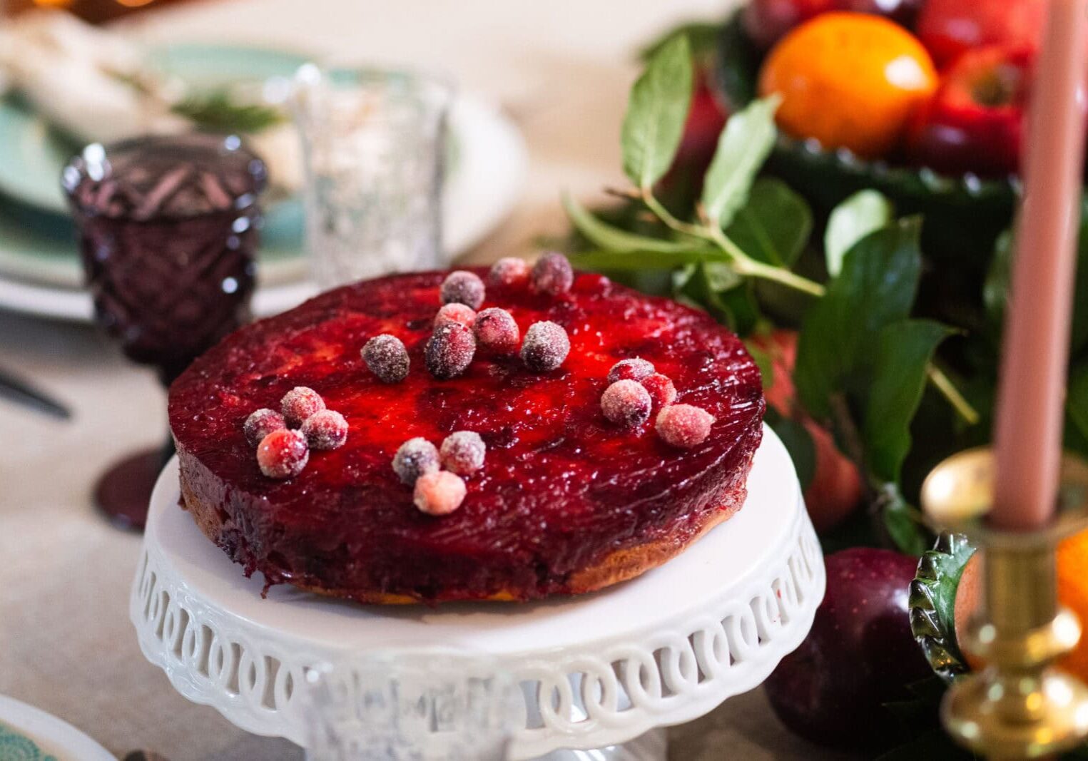 A round cranberry-topped cake on a white stand sits on a table set for a meal, with fruit and greenery in the background.