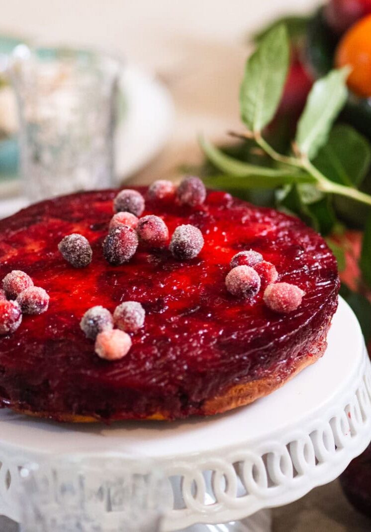 A round cranberry-topped cake on a white stand sits on a table set for a meal, with fruit and greenery in the background.