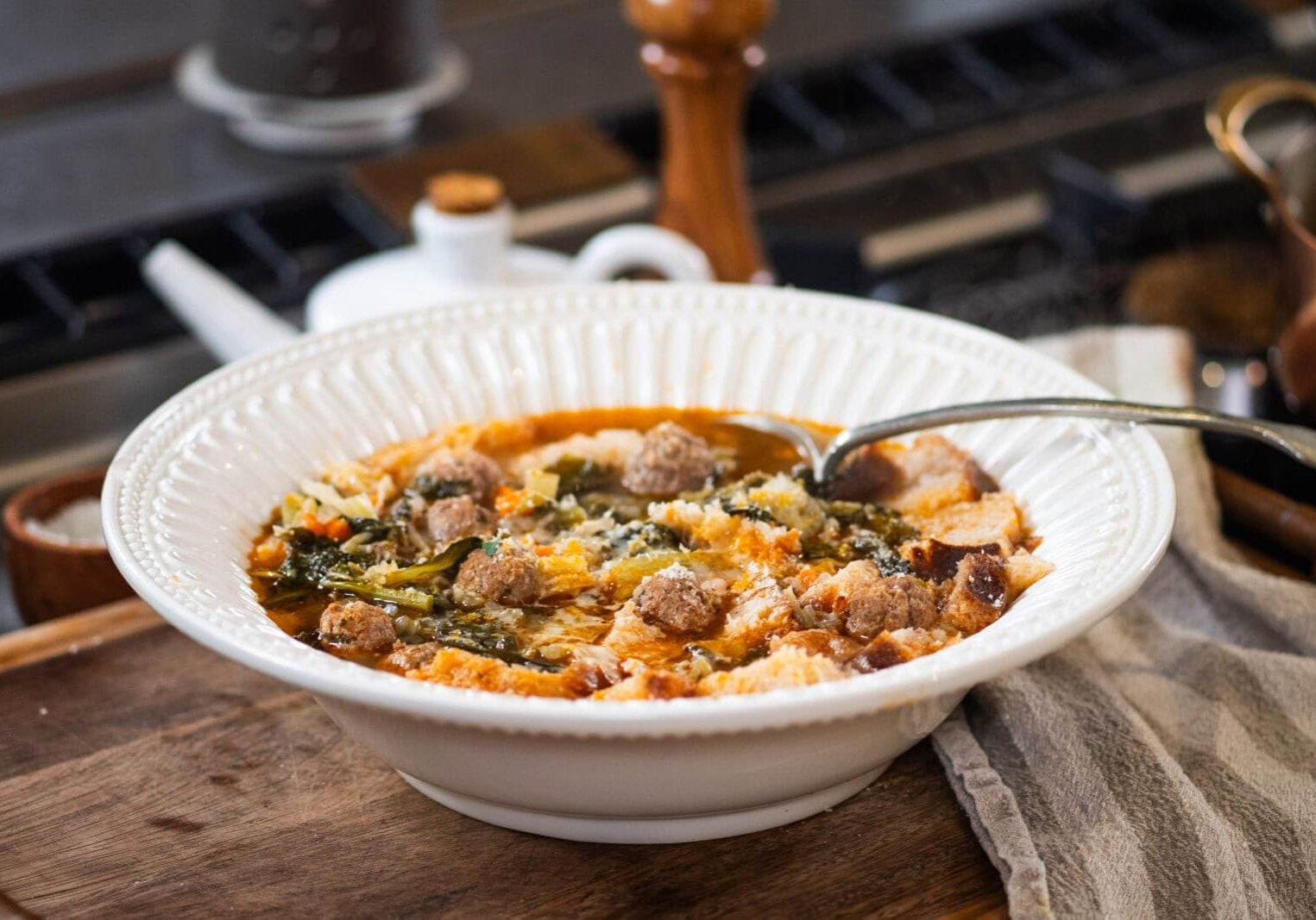 A white bowl filled with soup containing meatballs, vegetables, and bread pieces sits on a wooden surface next to a striped cloth napkin.