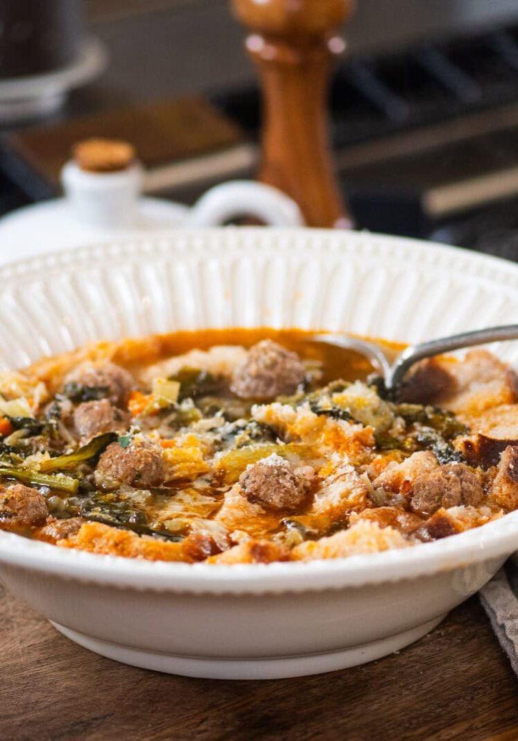 A white bowl filled with soup containing meatballs, vegetables, and bread pieces sits on a wooden surface next to a striped cloth napkin.