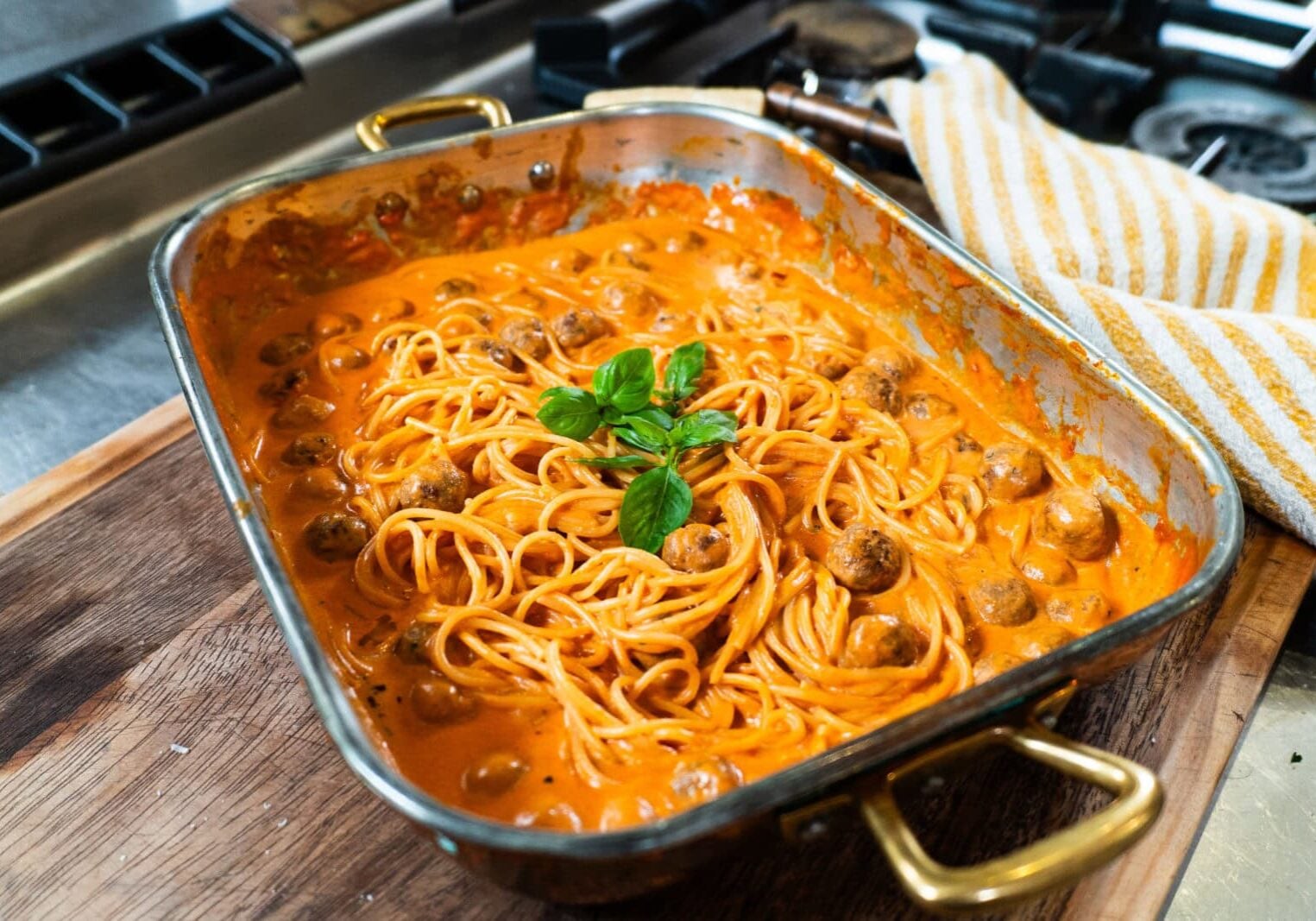 A baking dish filled with spaghetti and meatballs in a creamy tomato sauce, garnished with fresh basil, sits on a wooden board near a stove.