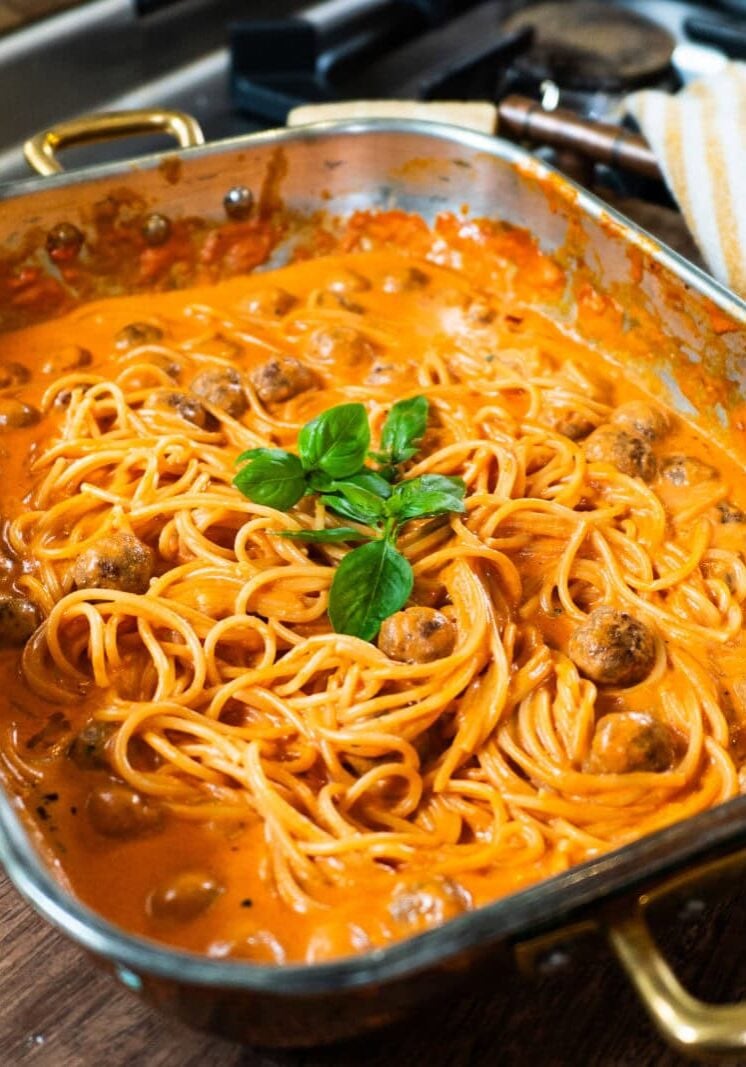A baking dish filled with spaghetti and meatballs in a creamy tomato sauce, garnished with fresh basil, sits on a wooden board near a stove.