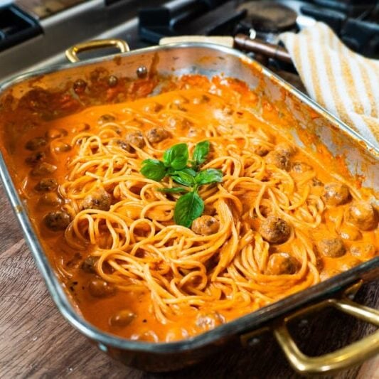 A baking dish filled with spaghetti and meatballs in a creamy tomato sauce, garnished with fresh basil, sits on a wooden board near a stove.