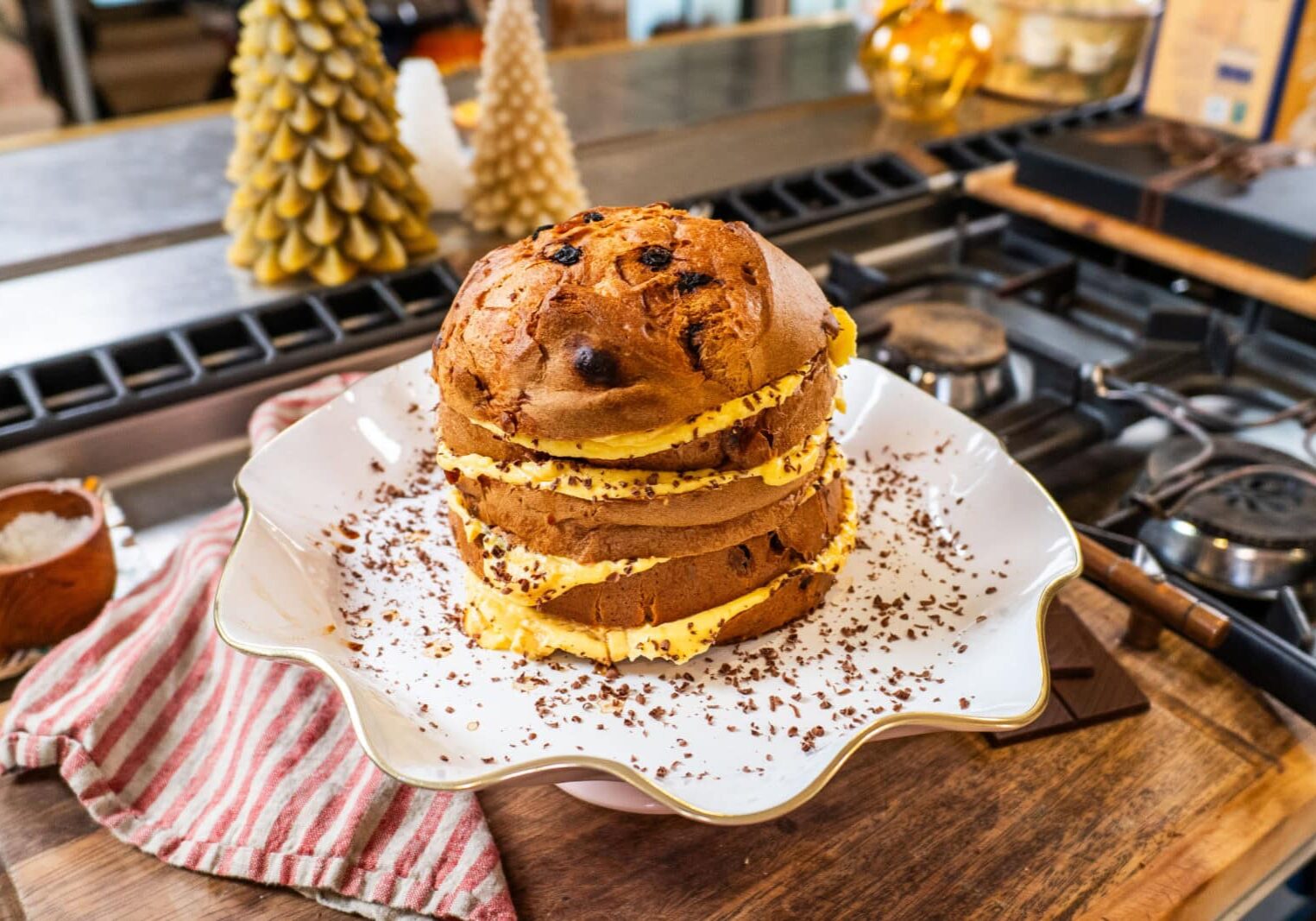 A sliced panettone cake with cream filling and chocolate shavings sits on a decorative plate on a kitchen counter.