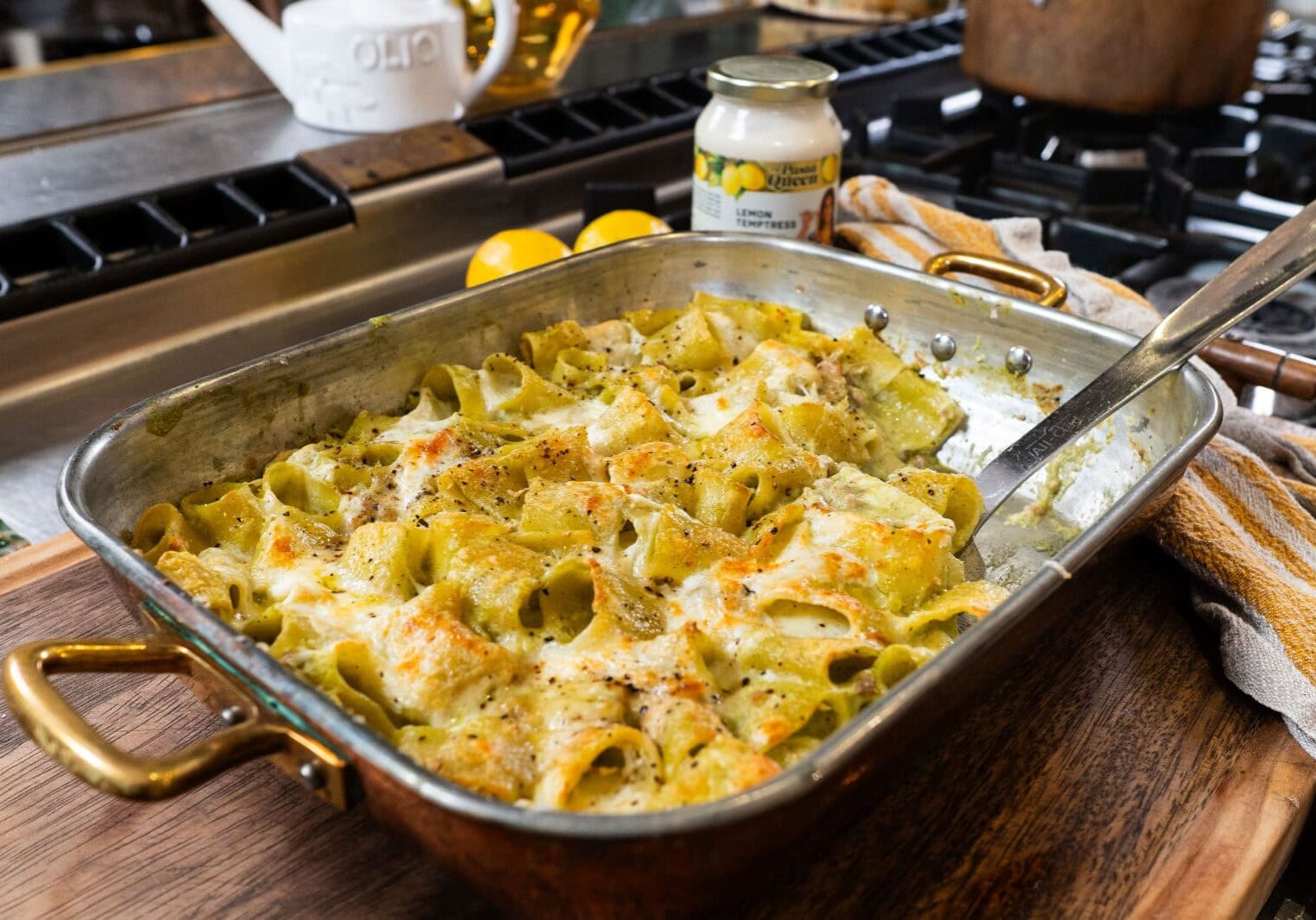 A baked pasta casserole with a creamy sauce is shown in a copper baking dish on a wooden board, with a serving utensil and a jar of lemon artichoke sauce nearby.