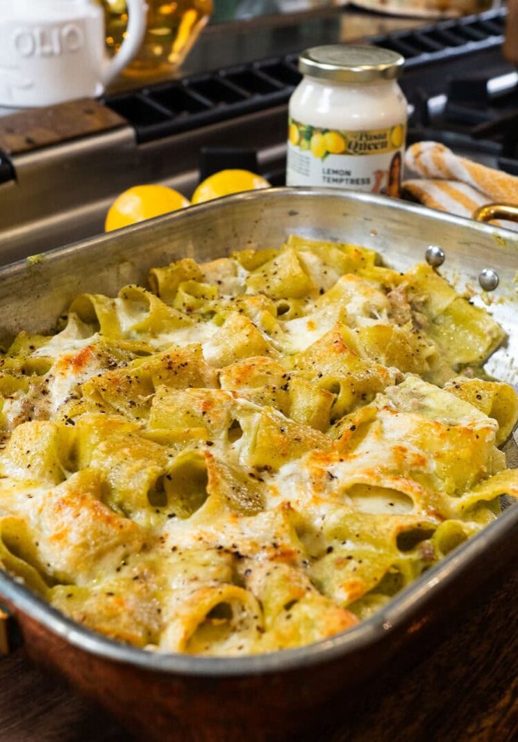 A baked pasta casserole with a creamy sauce is shown in a copper baking dish on a wooden board, with a serving utensil and a jar of lemon artichoke sauce nearby.