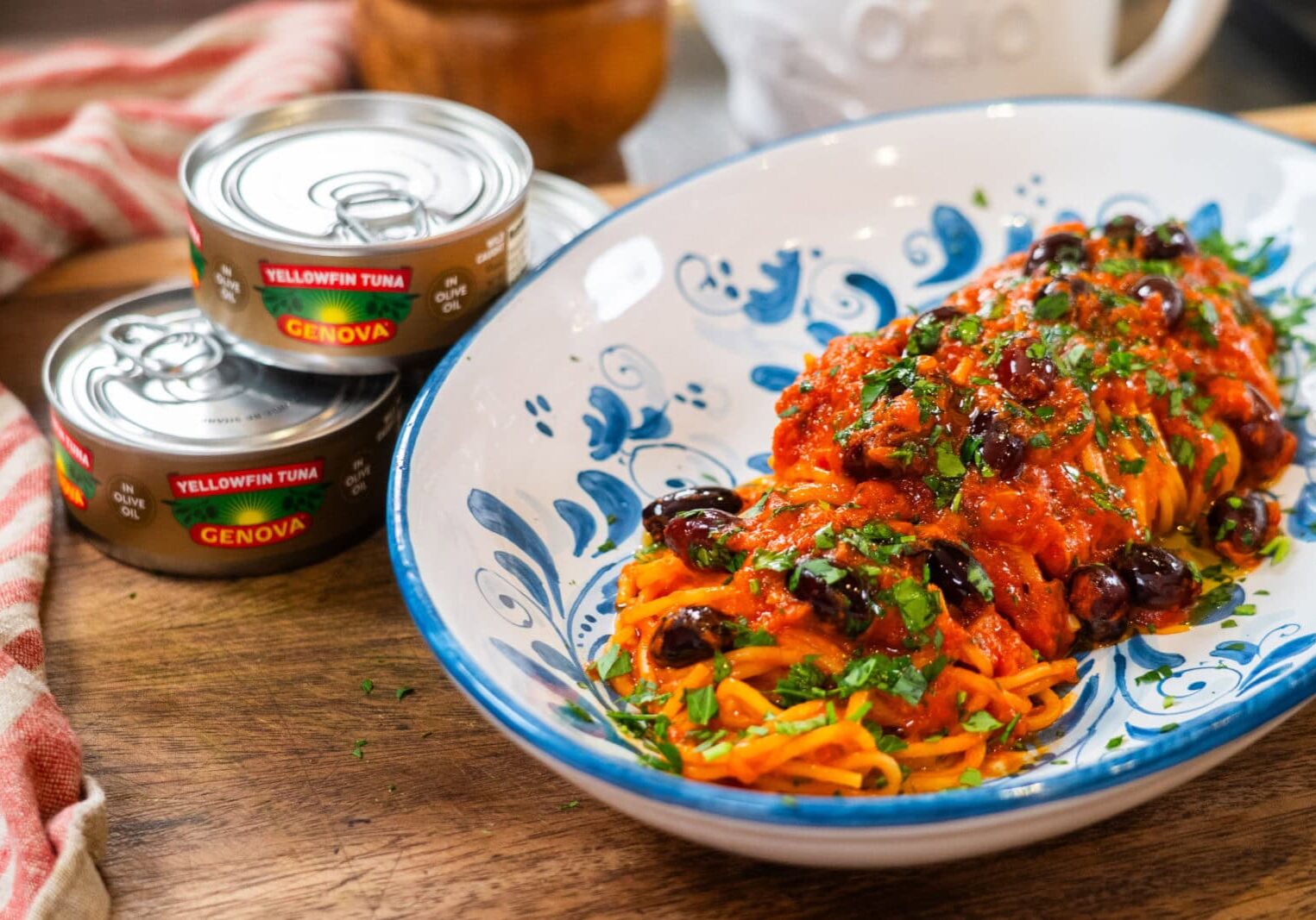 A plate of spaghetti topped with tomato sauce, black olives, and herbs sits beside two cans of Genova yellowfin tuna on a wooden surface.
