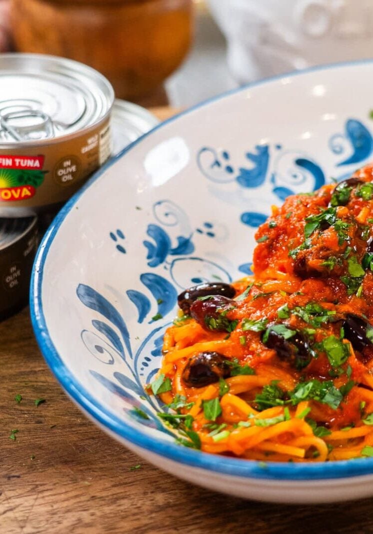 A plate of spaghetti topped with tomato sauce, black olives, and herbs sits beside two cans of Genova yellowfin tuna on a wooden surface.