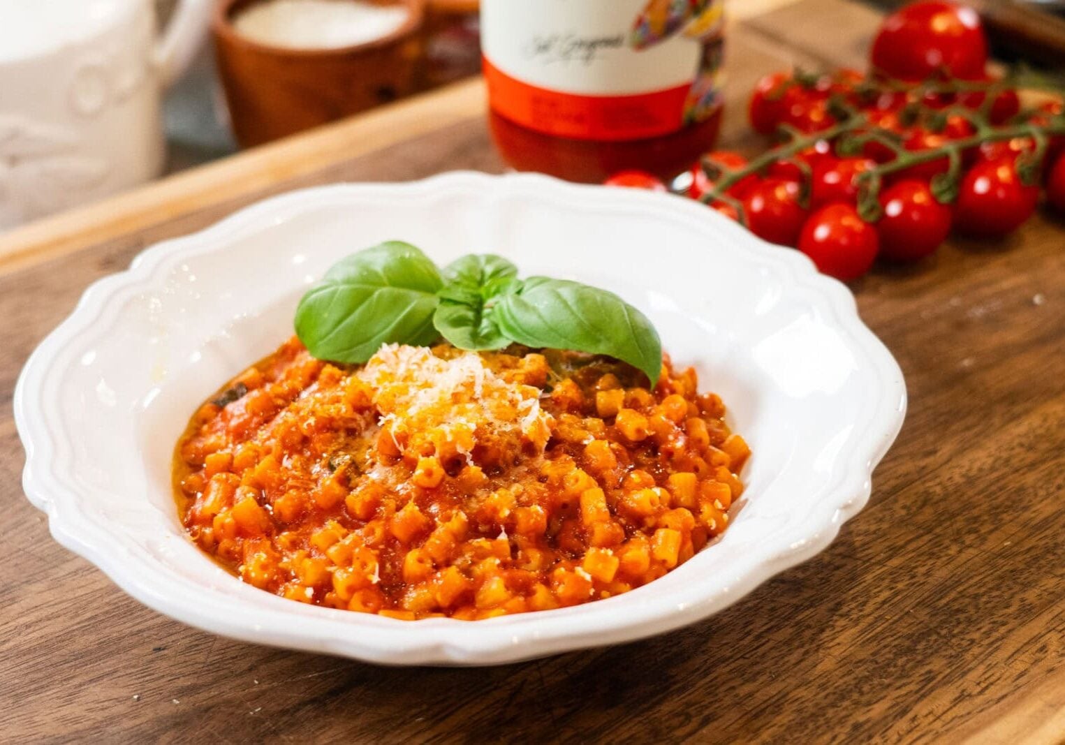 A white bowl of tomato-based pasta garnished with basil and grated cheese sits on a wooden table, with cherry tomatoes and a sauce bottle in the background.