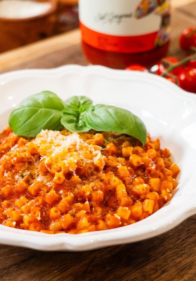 A white bowl of tomato-based pasta garnished with basil and grated cheese sits on a wooden table, with cherry tomatoes and a sauce bottle in the background.