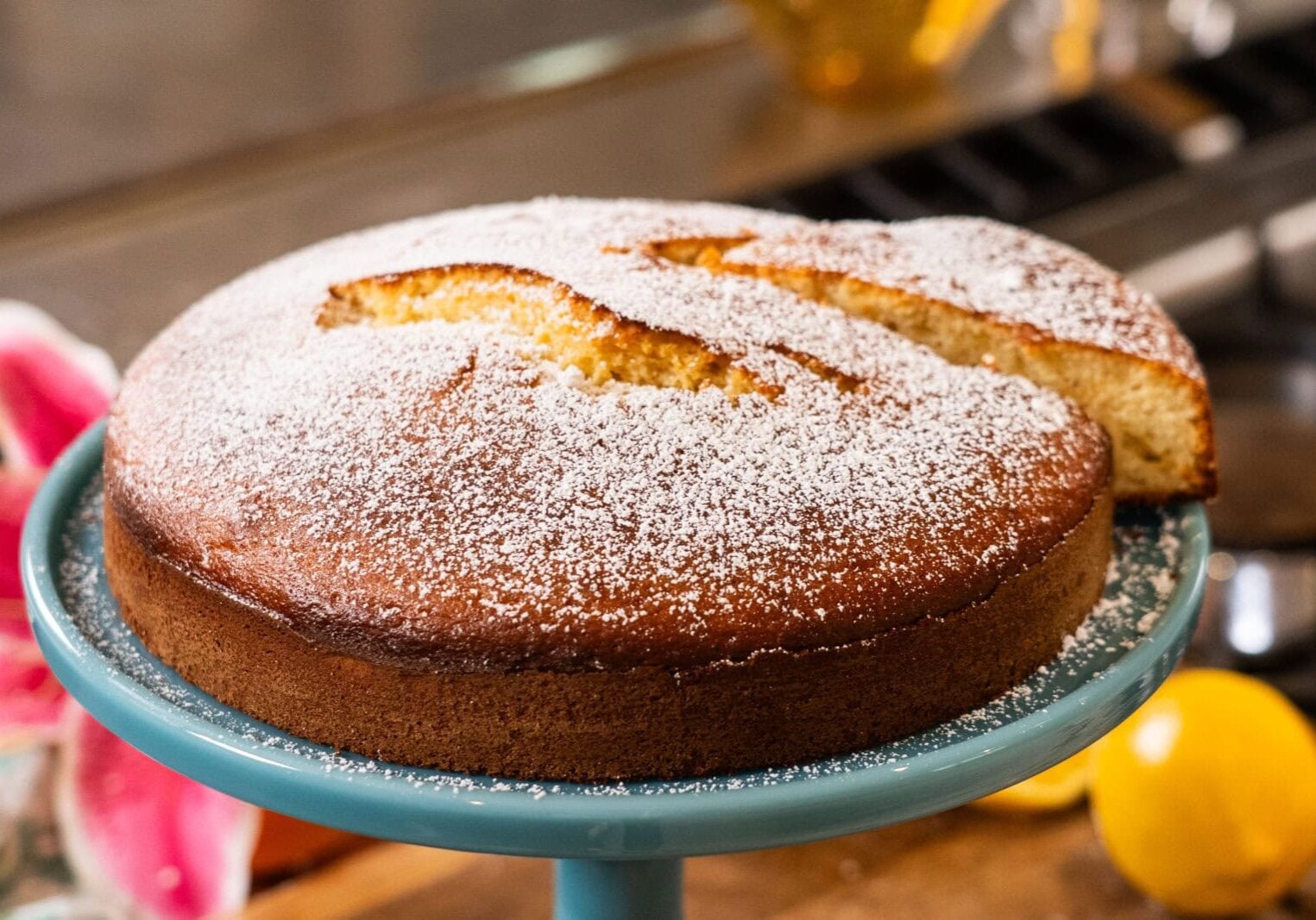 A round, golden-brown cake dusted with powdered sugar sits on a blue cake stand, with one slice cut and slightly separated from the whole cake.
