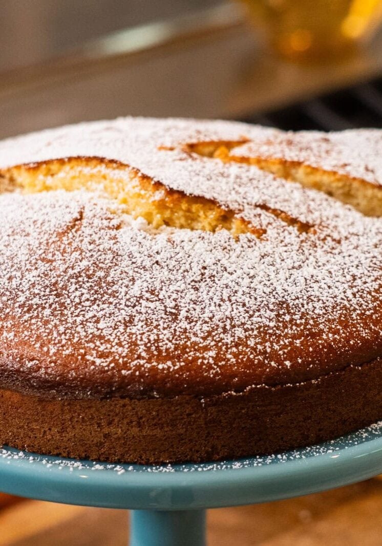 A round, golden-brown cake dusted with powdered sugar sits on a blue cake stand, with one slice cut and slightly separated from the whole cake.