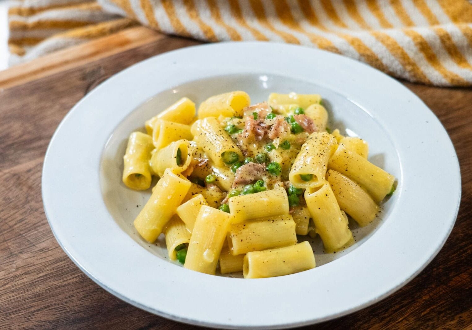 A white plate of rigatoni pasta with peas, ground meat, and a creamy sauce, garnished with black pepper, on a wooden surface.