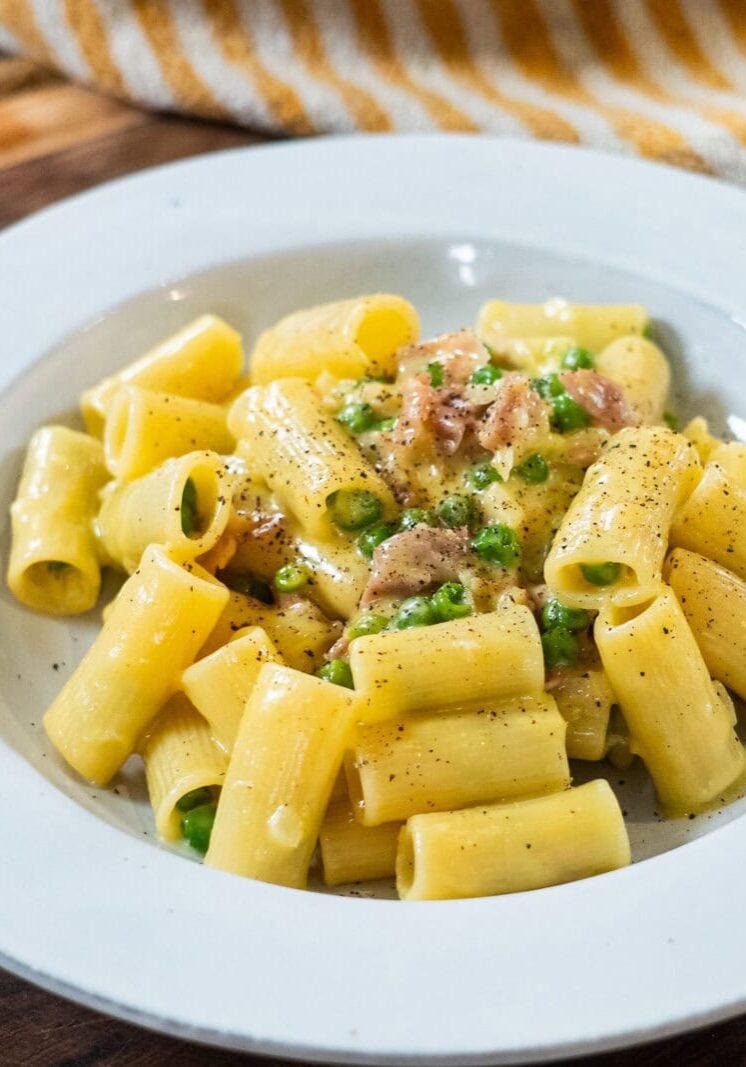 A white plate of rigatoni pasta with peas, ground meat, and a creamy sauce, garnished with black pepper, on a wooden surface.