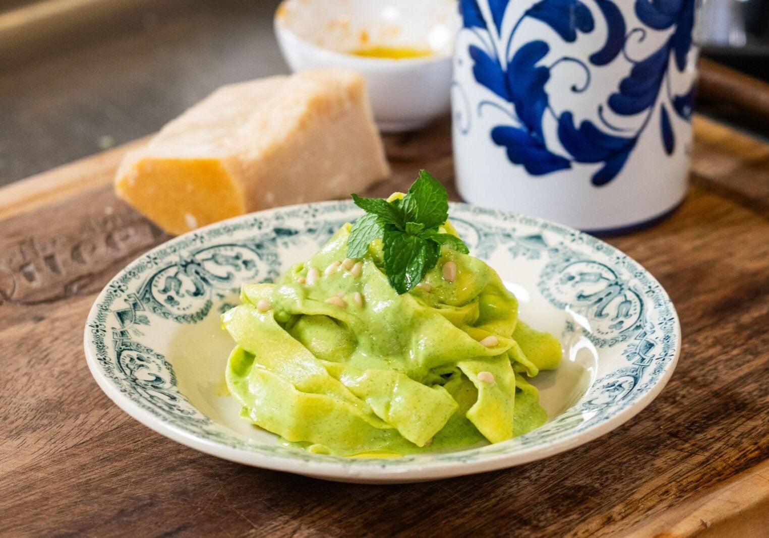 A plate of pasta with green sauce is garnished with mint leaves on a wooden surface, alongside a block of cheese and a blue-and-white container.