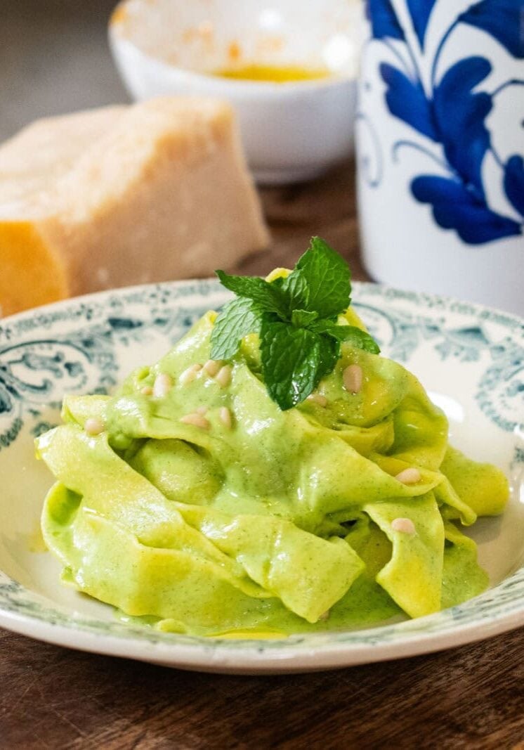A plate of pasta with green sauce is garnished with mint leaves on a wooden surface, alongside a block of cheese and a blue-and-white container.