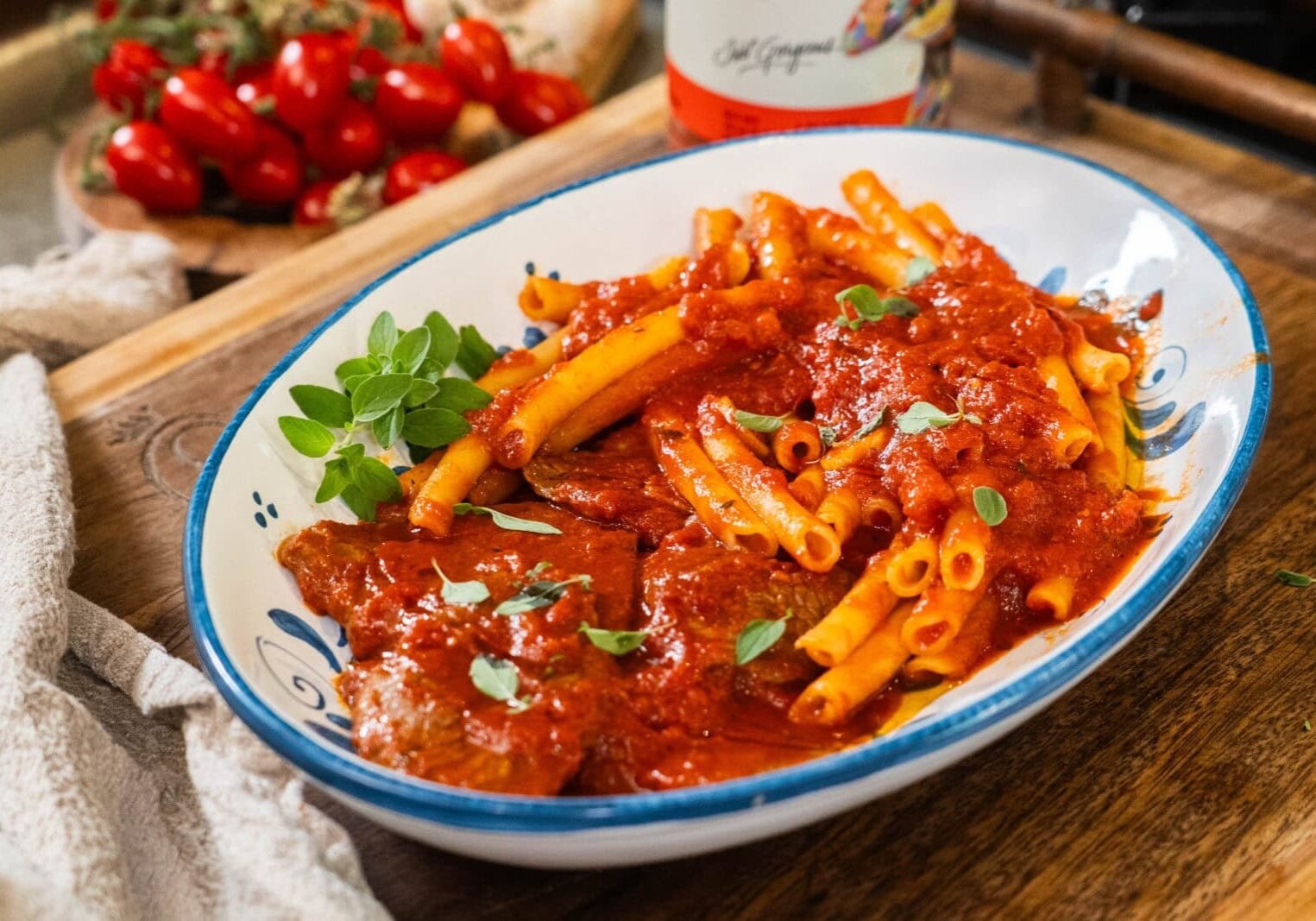 A white oval dish with ziti pasta and meat covered in tomato sauce, garnished with fresh herbs, sits on a wooden surface.