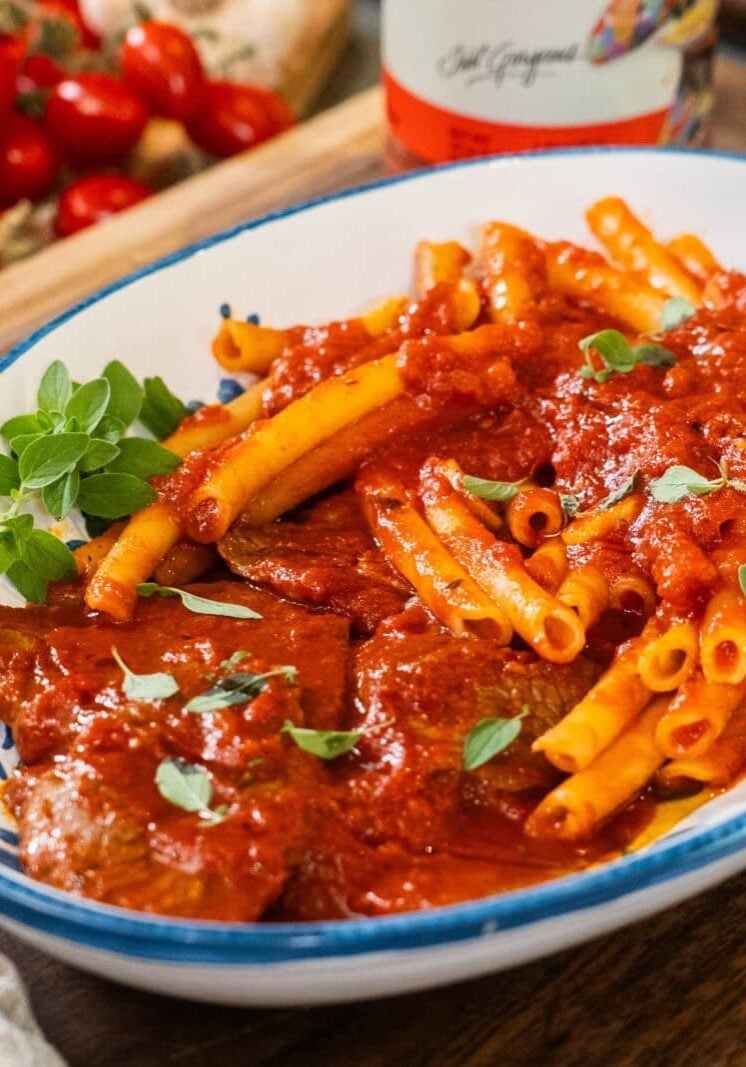 A white oval dish with ziti pasta and meat covered in tomato sauce, garnished with fresh herbs, sits on a wooden surface.