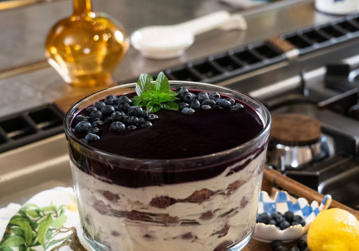 A layered trifle dessert with cream, cake, and blueberry topping garnished with mint leaves, sitting on a kitchen counter near a bowl of fresh blueberries.