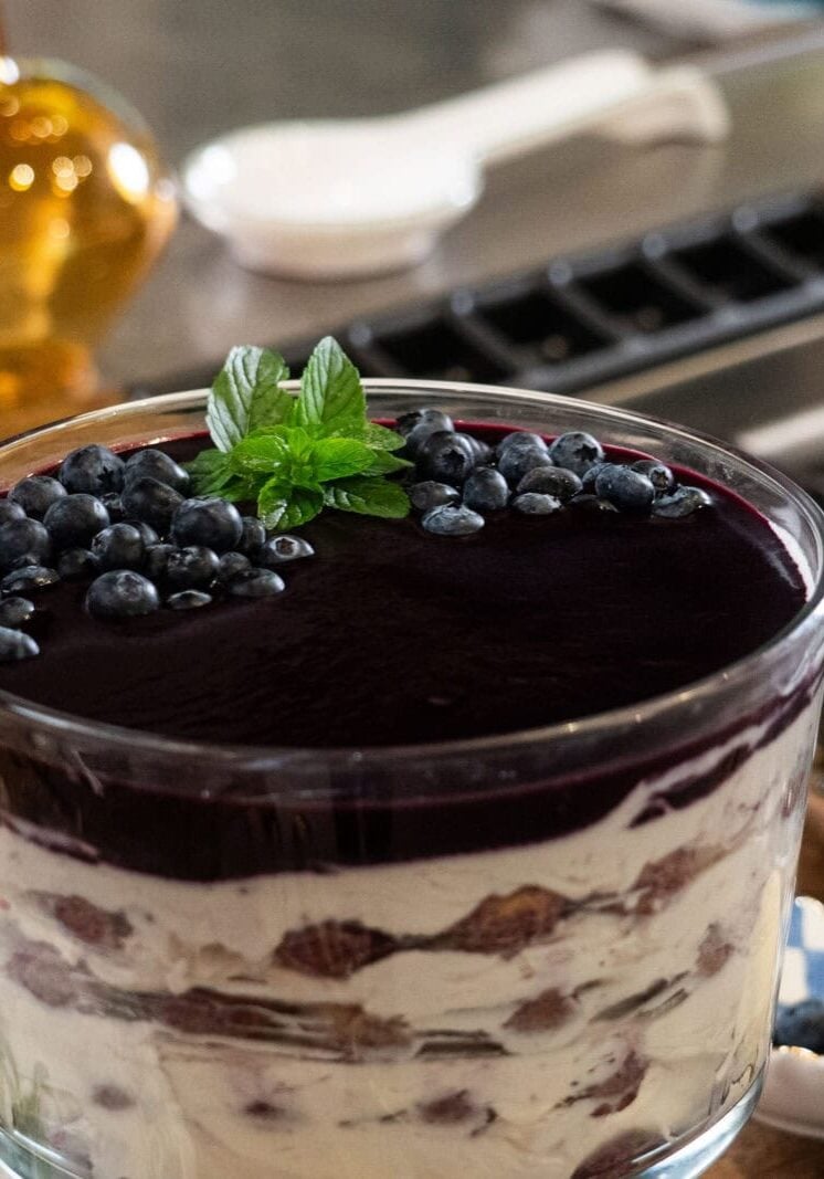 A layered trifle dessert with cream, cake, and blueberry topping garnished with mint leaves, sitting on a kitchen counter near a bowl of fresh blueberries.