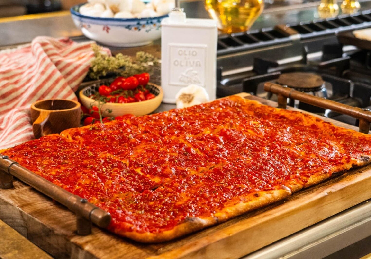 Rectangular sheet pan pizza with tomato sauce sits on a wooden board in a kitchen, surrounded by ingredients like garlic, tomatoes, and olive oil.