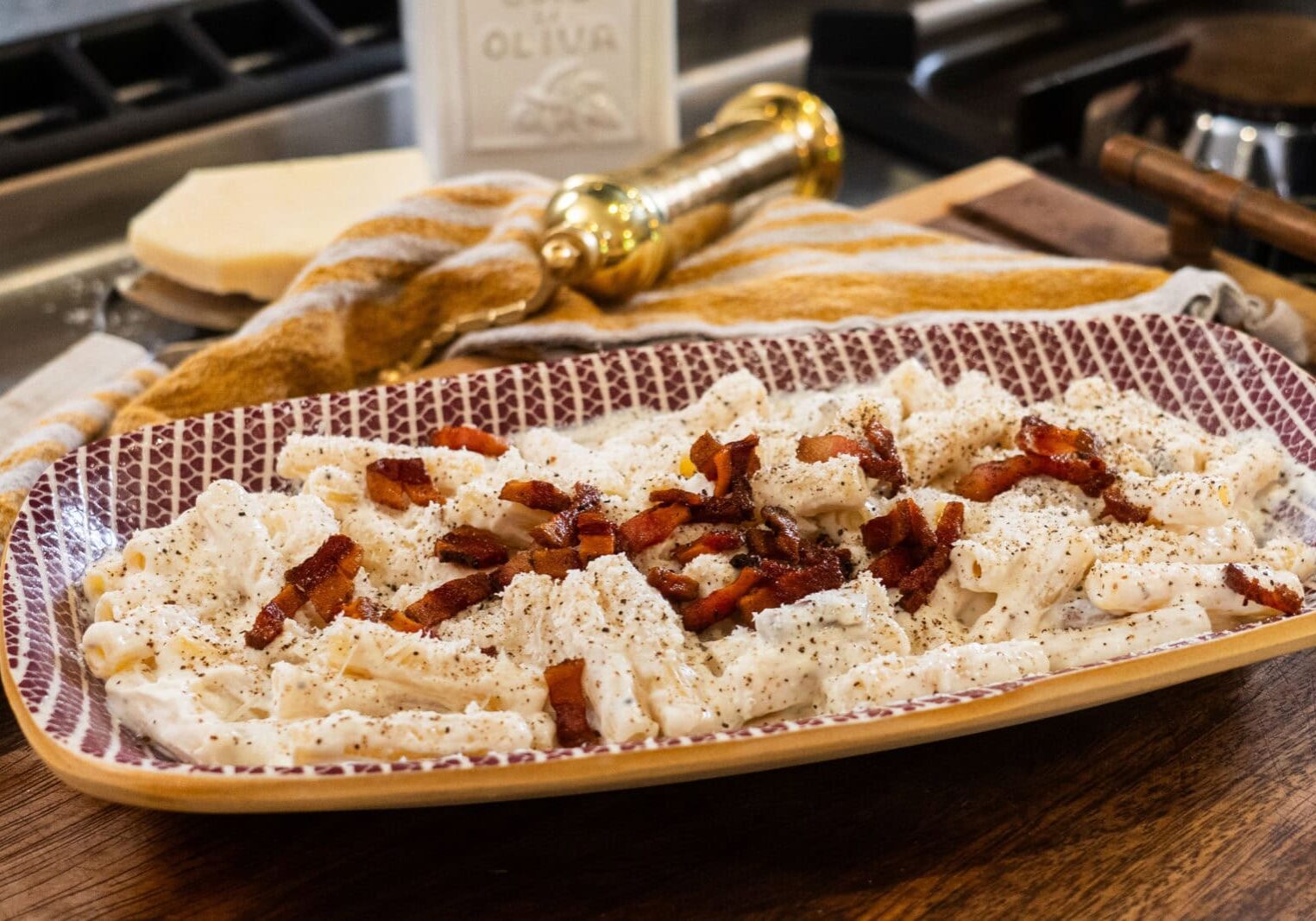 A rectangular plate of creamy pasta topped with crumbled bacon and black pepper is on a wooden surface, with a cheese wedge, a gold grinder, and a striped towel in the background.