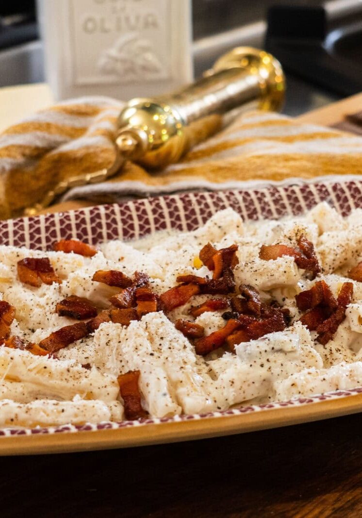 A rectangular plate of creamy pasta topped with crumbled bacon and black pepper is on a wooden surface, with a cheese wedge, a gold grinder, and a striped towel in the background.