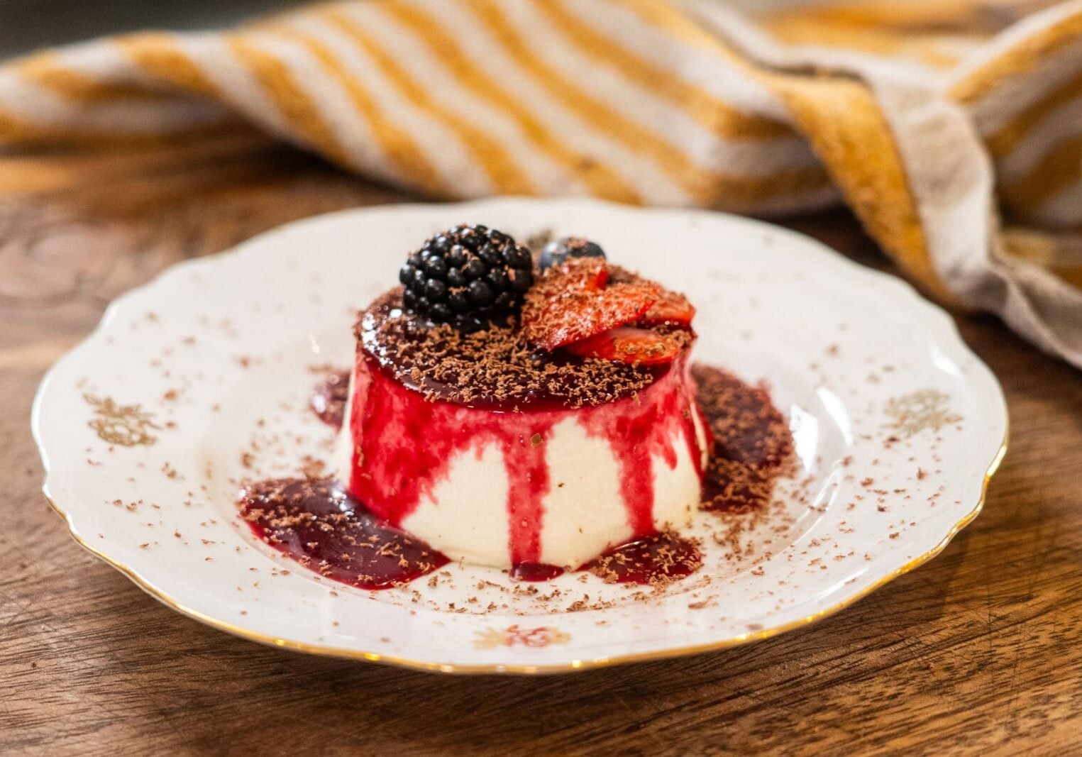 A dessert of panna cotta with berry sauce, topped with sliced strawberries, a blackberry, and chocolate shavings, served on a decorative white plate.