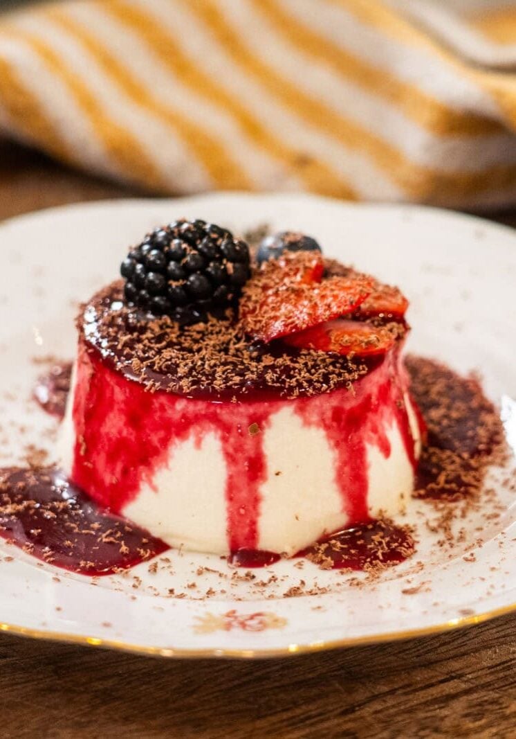 A dessert of panna cotta with berry sauce, topped with sliced strawberries, a blackberry, and chocolate shavings, served on a decorative white plate.