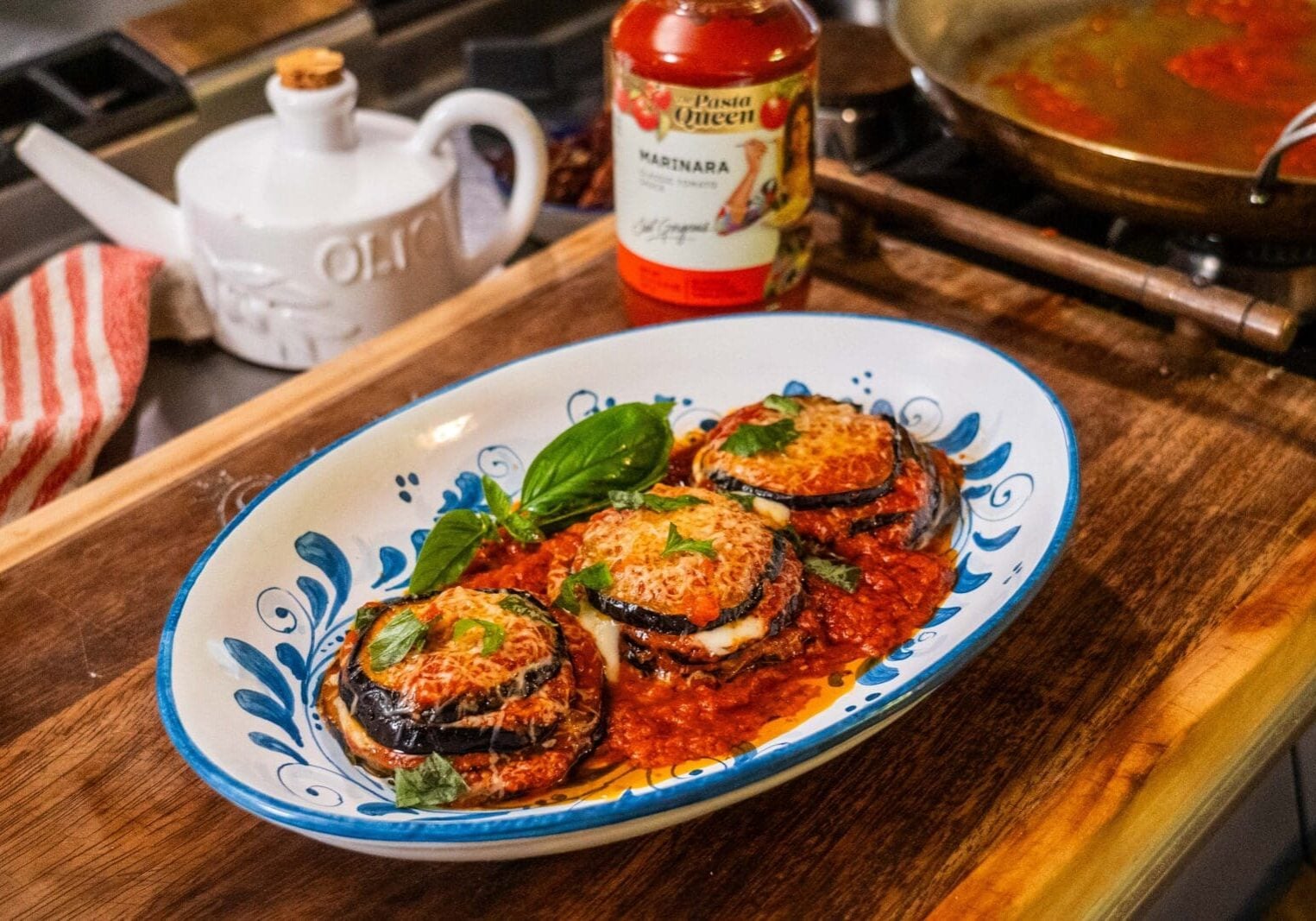 Three baked eggplant parmesan stacks with marinara sauce and melted cheese, garnished with basil, served on a decorative plate next to a jar of marinara sauce and a cooking pan.