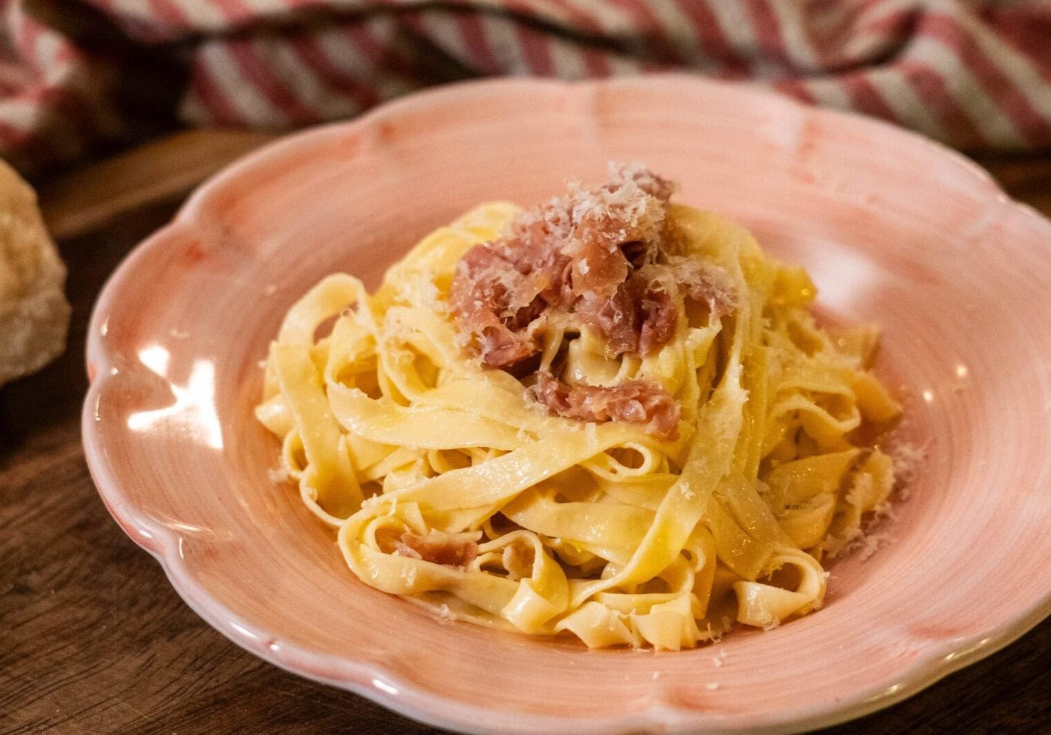 A plate of tagliatelle pasta topped with grated cheese and pieces of ham sits on a pink dish with a striped cloth in the background.