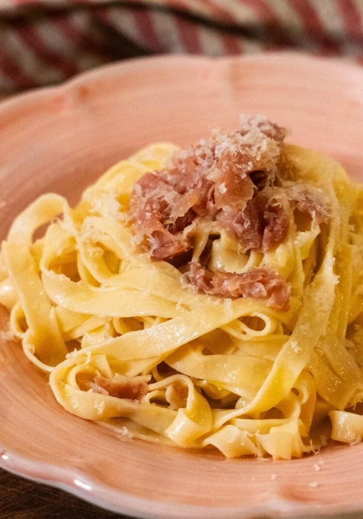 A plate of tagliatelle pasta topped with grated cheese and pieces of ham sits on a pink dish with a striped cloth in the background.