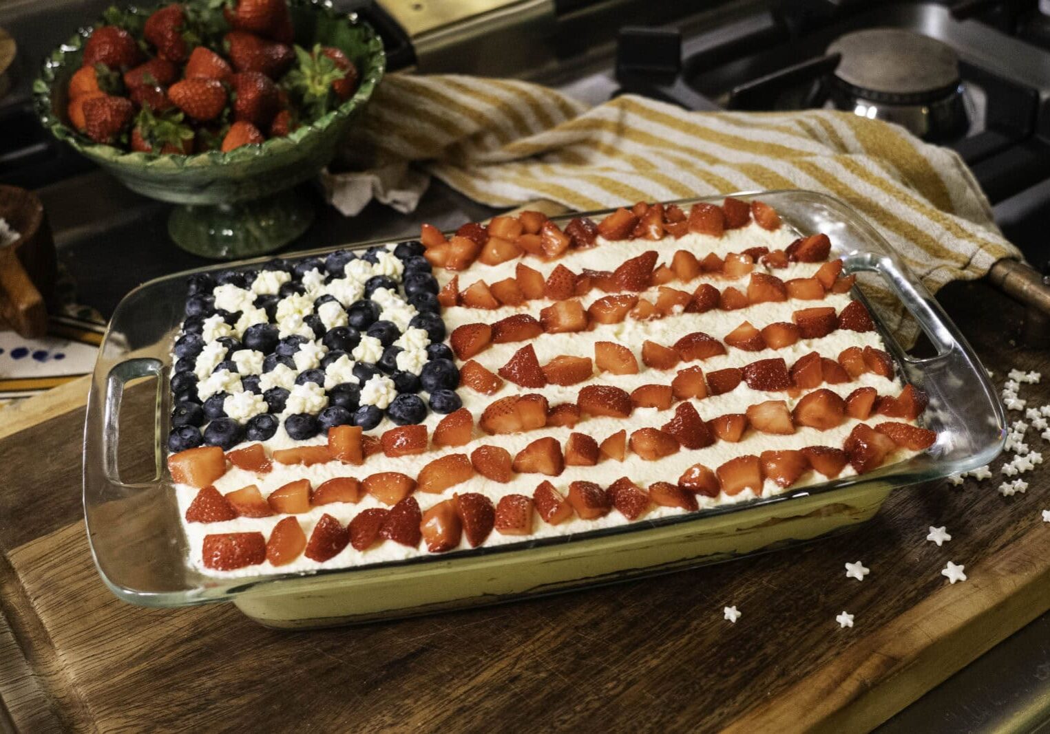 A rectangular dessert decorated like the American flag with blueberries, strawberries, and white cream sits on a wooden surface near a bowl of strawberries and a striped towel.