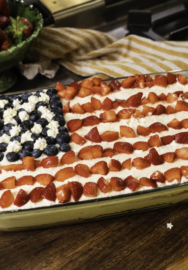 A rectangular dessert decorated like the American flag with blueberries, strawberries, and white cream sits on a wooden surface near a bowl of strawberries and a striped towel.