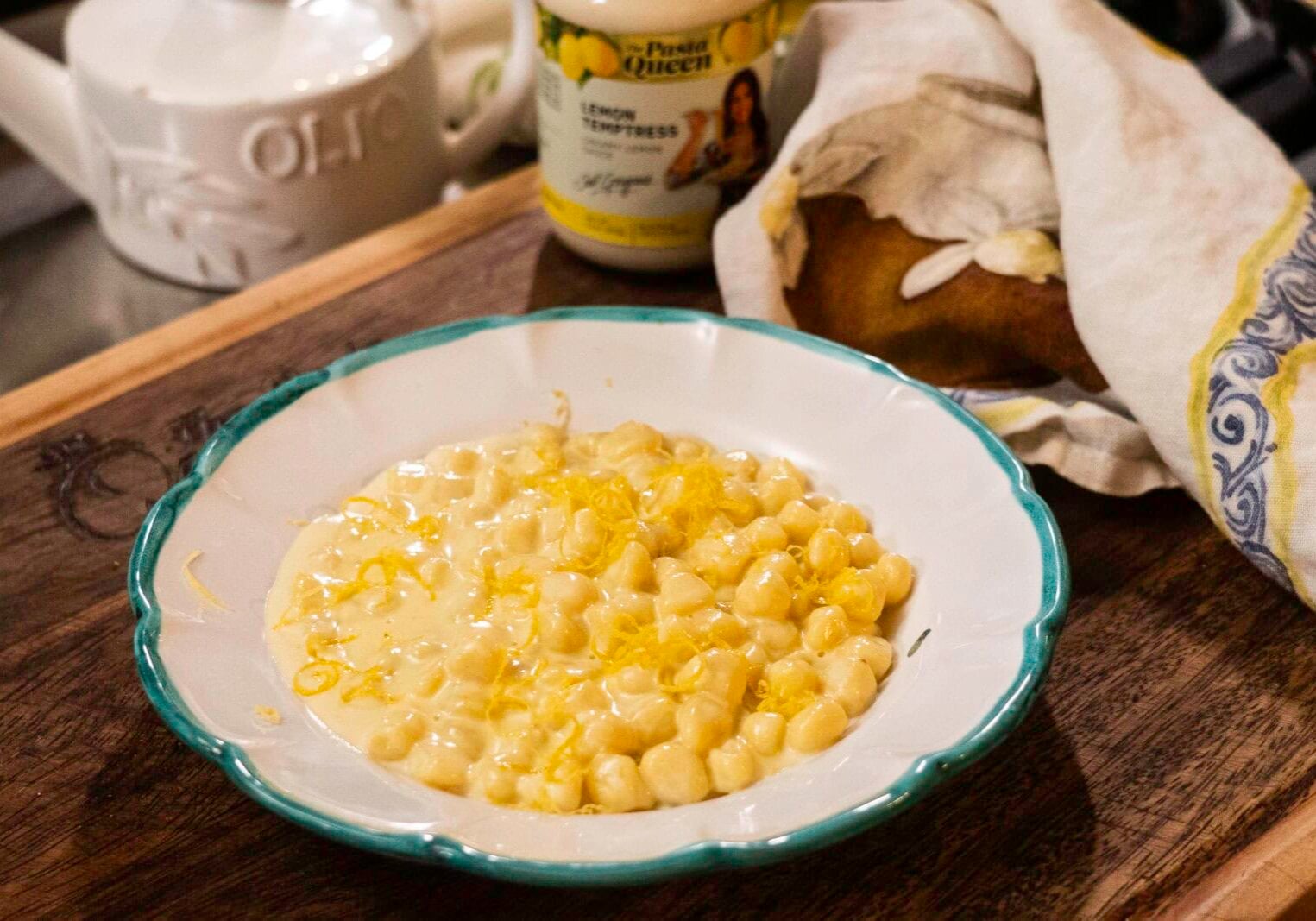 A white bowl with green trim contains creamy pasta shells topped with lemon zest, set on a wooden surface beside a loaf of bread and a jar labeled "Lemon Empress.