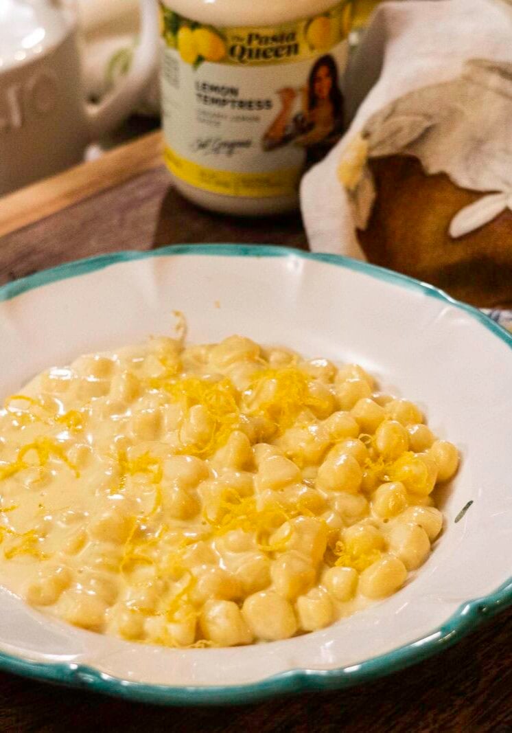 A white bowl with green trim contains creamy pasta shells topped with lemon zest, set on a wooden surface beside a loaf of bread and a jar labeled "Lemon Empress.