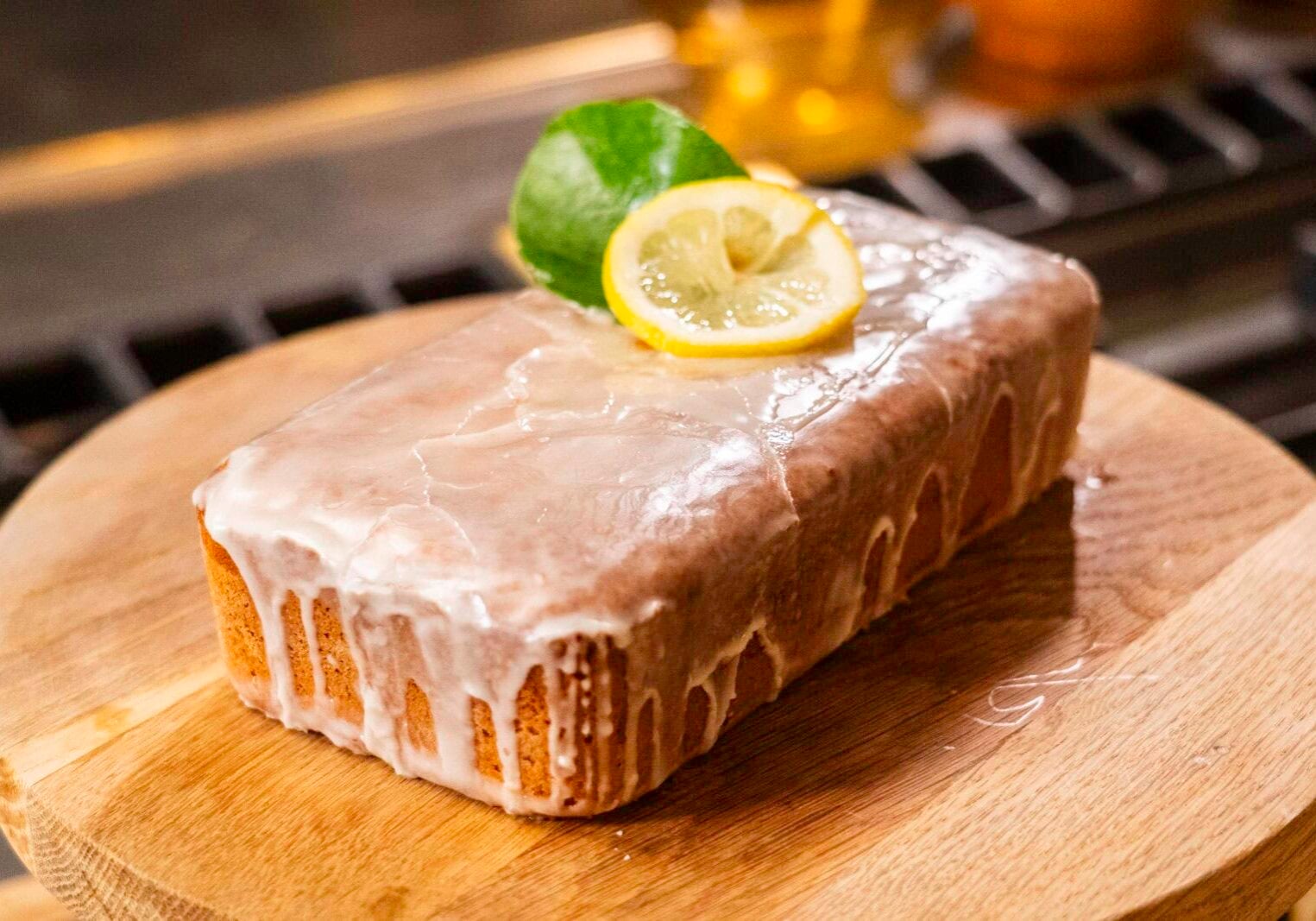 A glazed loaf cake topped with a lemon slice and green leaf sits on a wooden board.