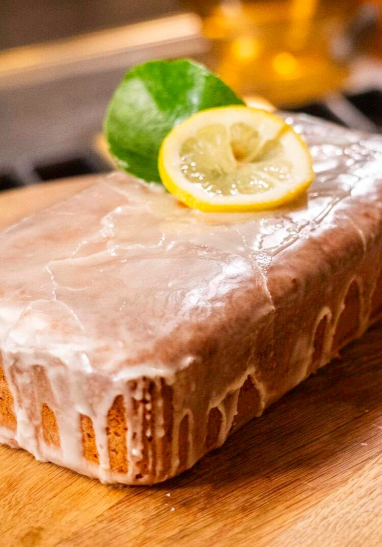 A glazed loaf cake topped with a lemon slice and green leaf sits on a wooden board.