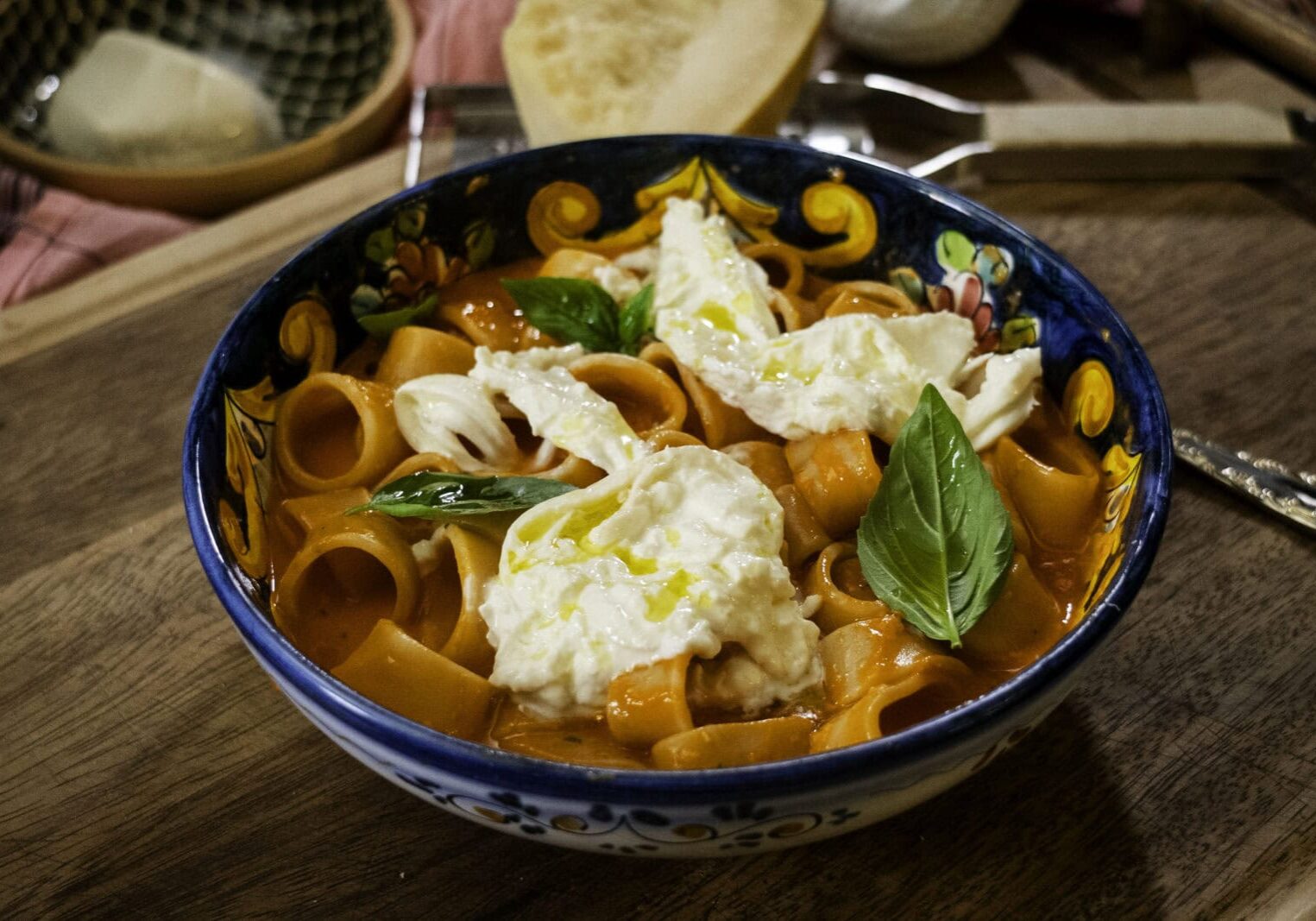 A bowl of pasta in tomato sauce topped with burrata cheese and fresh basil sits on a wooden surface beside a slice of bread.
