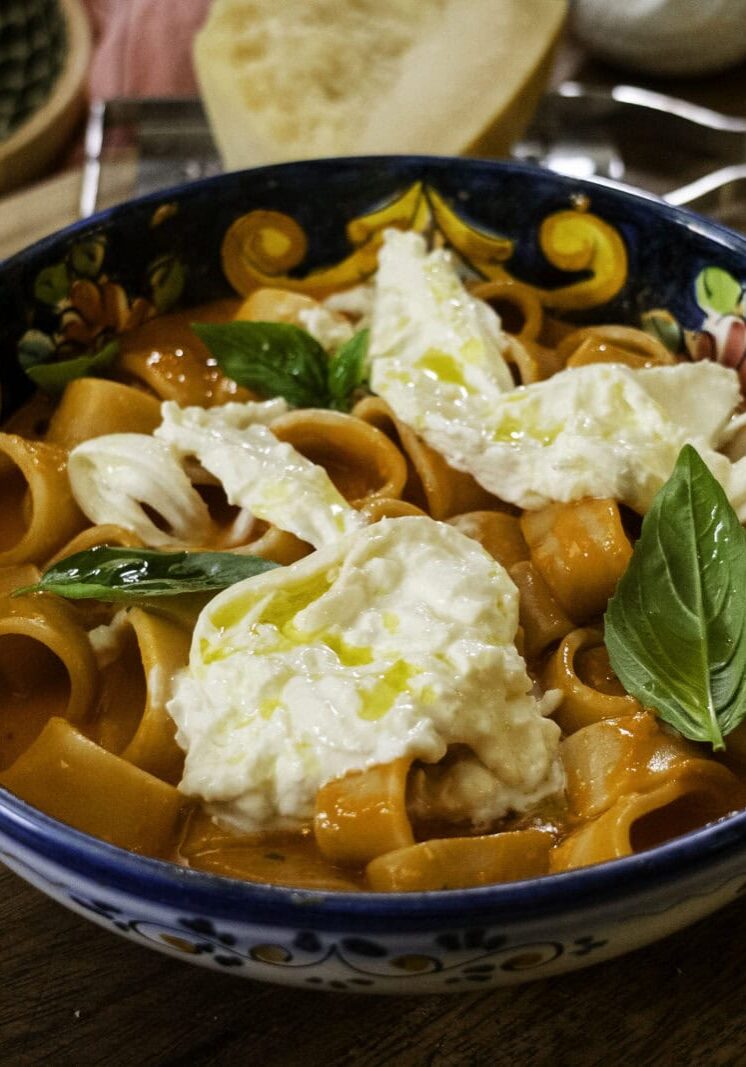 A bowl of pasta in tomato sauce topped with burrata cheese and fresh basil sits on a wooden surface beside a slice of bread.