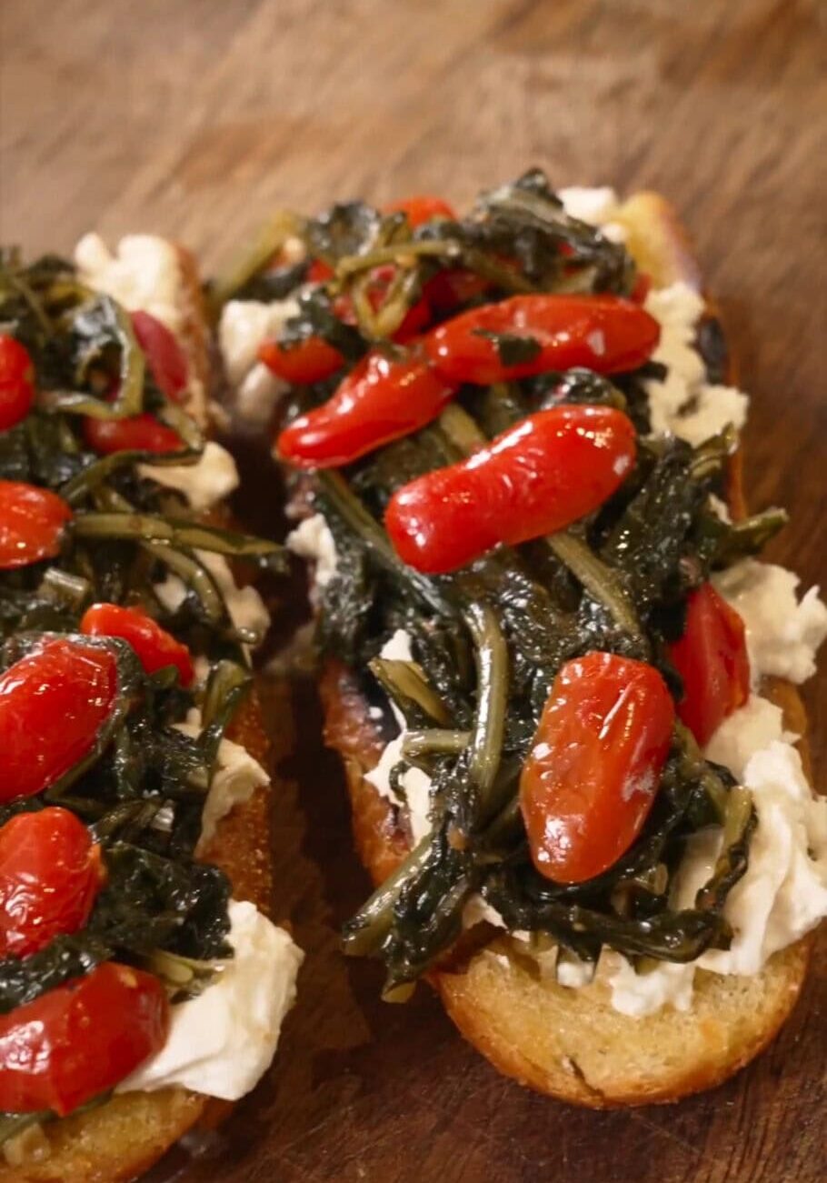 Two slices of toast topped with cream cheese, sautéed greens, and cherry tomatoes.