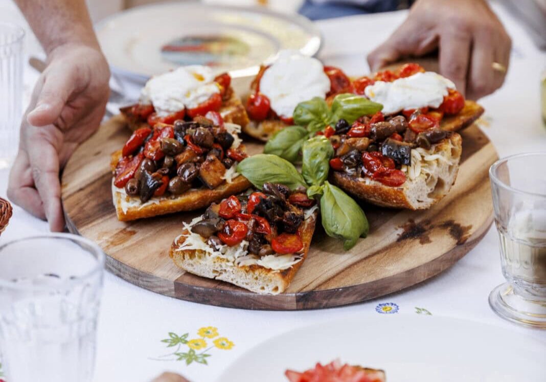 Hands placing a large wooden board with bruschetta topped with tomatoes, basil, and mozzarella on a table. A person reaches for a slice.