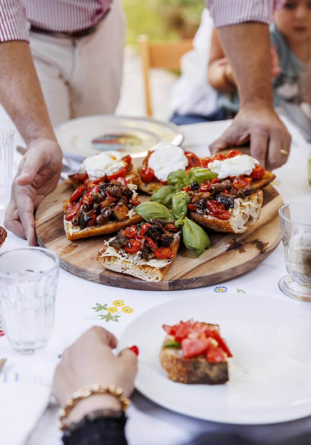 Hands placing a large wooden board with bruschetta topped with tomatoes, basil, and mozzarella on a table. A person reaches for a slice.