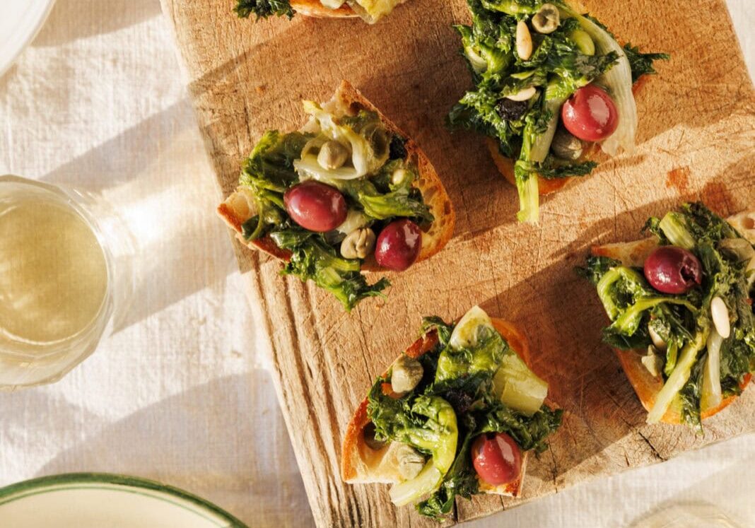 Slices of bread topped with greens, red grapes, and seeds on a wooden board, with glasses of white wine and plates nearby.