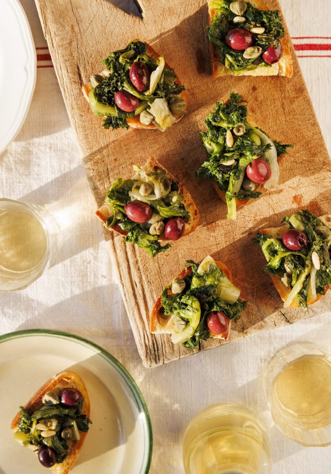 Slices of bread topped with greens, red grapes, and seeds on a wooden board, with glasses of white wine and plates nearby.