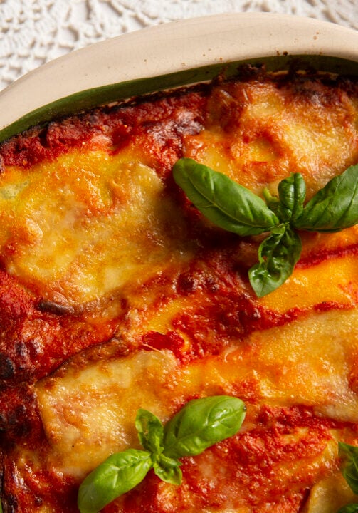 A baked dish of lasagna topped with melted cheese and garnished with fresh basil leaves, placed on a white lace tablecloth.