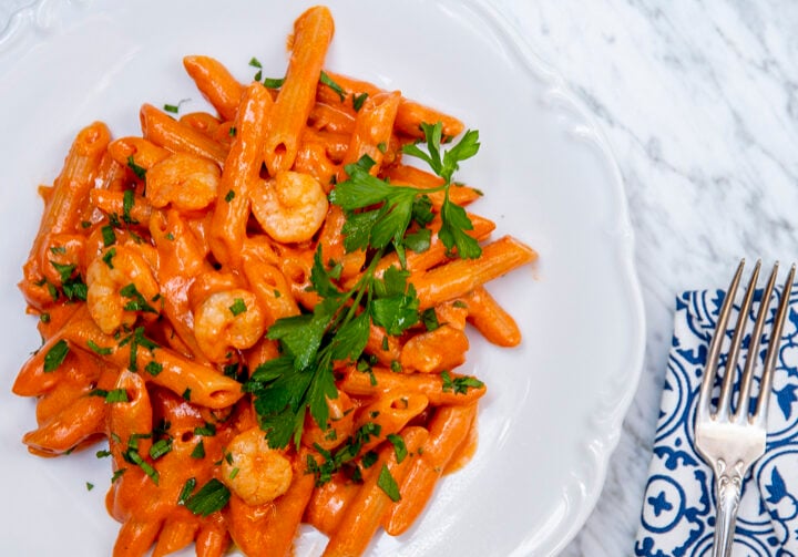 Plate of creamy penne pasta with shrimp, garnished with parsley. Placed on a marble surface near a patterned napkin and fork.