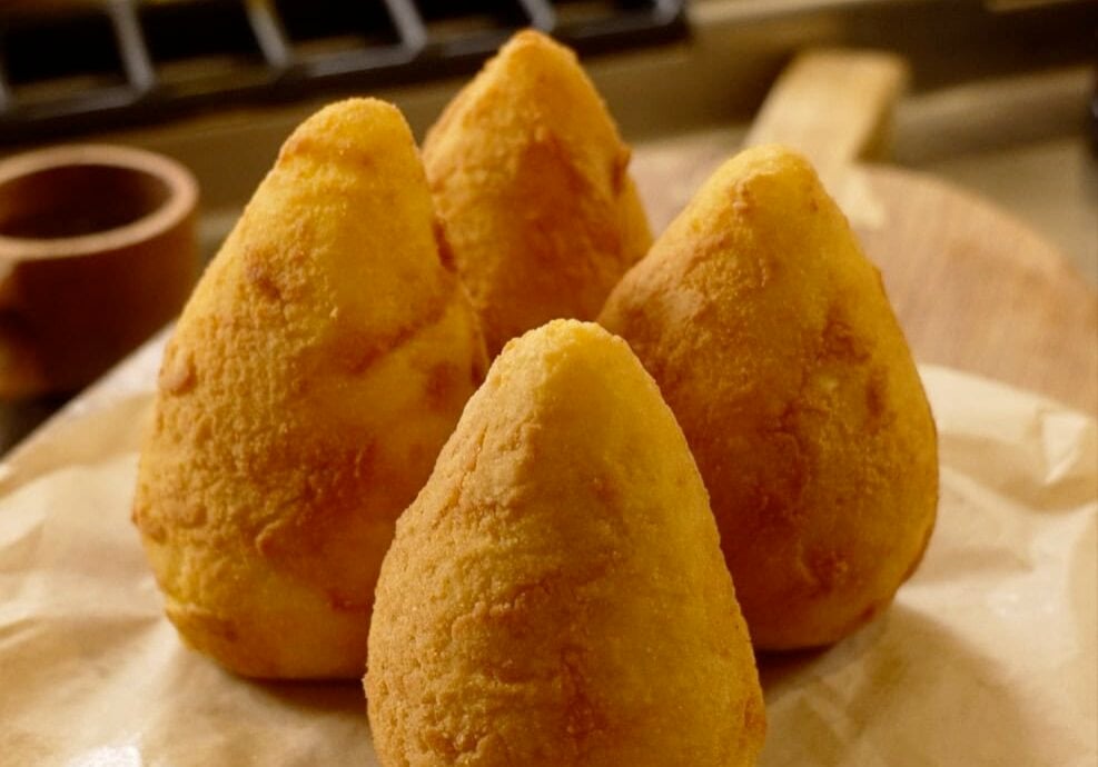 Four golden, cone-shaped fried rice balls sit on parchment paper, placed on a wooden board in a kitchen setting.